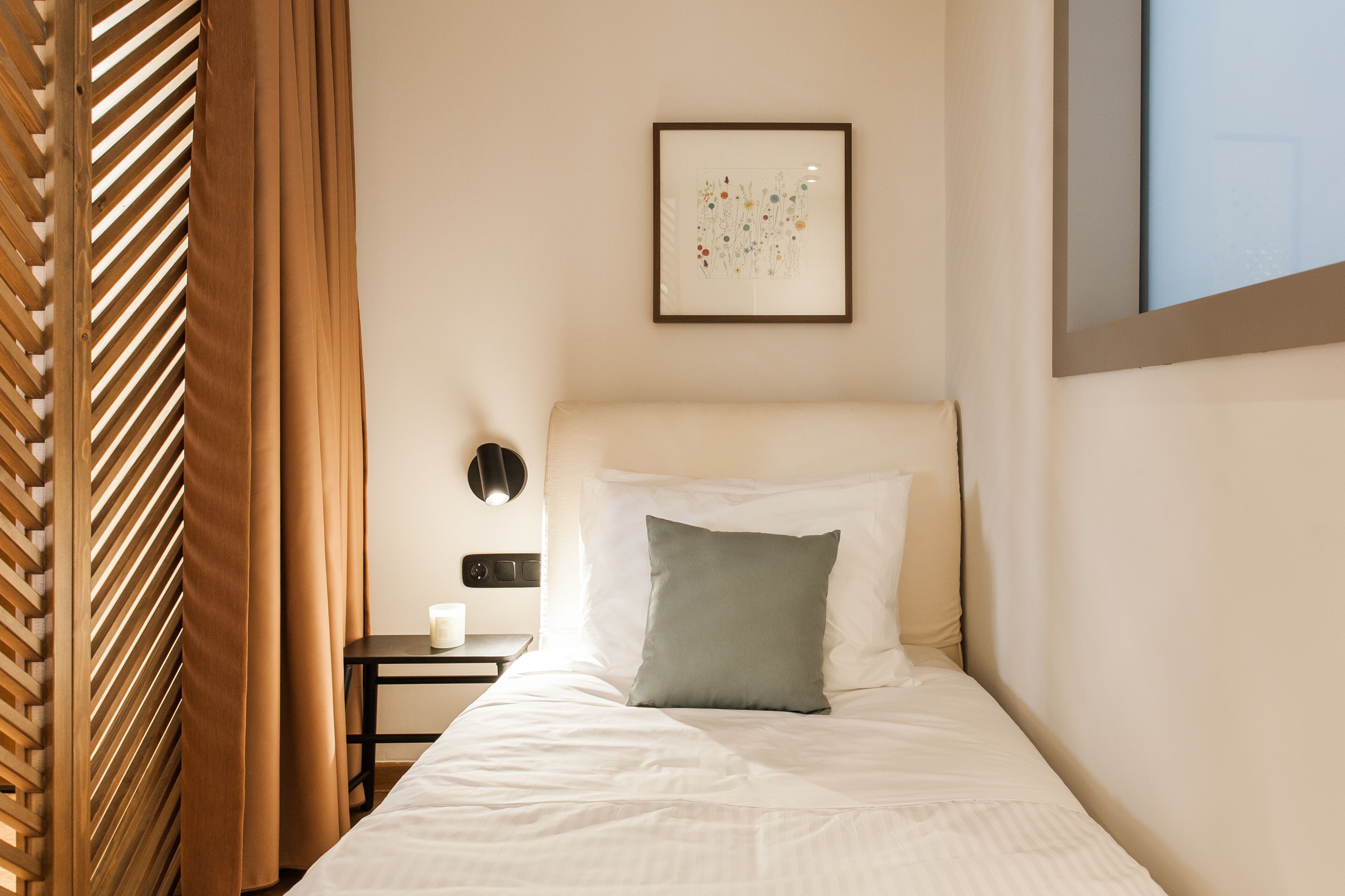 Una cama bien hecha con sábanas blancas y una única almohada verde, una mesita de noche con lámpara, y una obra de arte floral enmarcada en la pared.