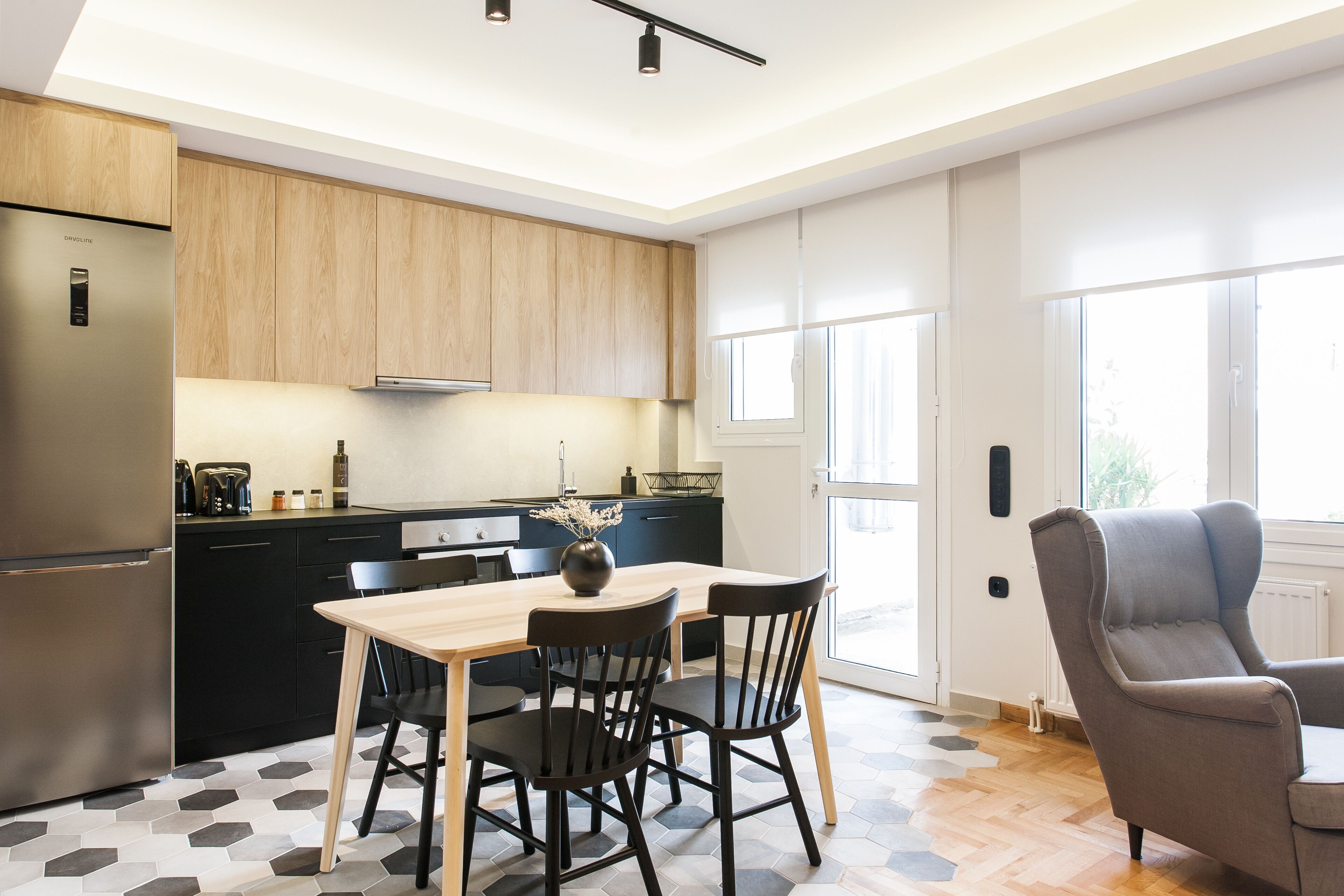 A modern kitchen with a built-in refrigerator, wooden cabinets, a dining table with chairs, and a large window providing natural light.