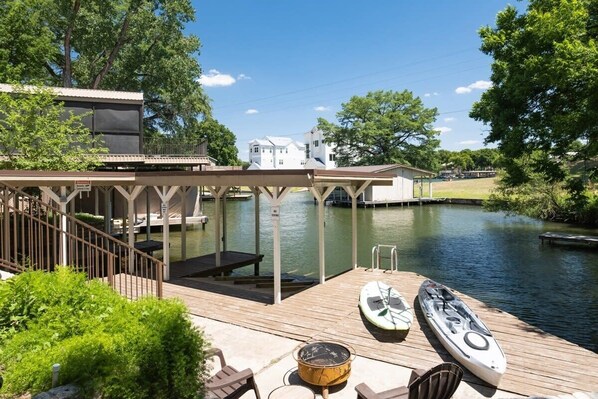 Un muelle con kayaks, una zona de estar cubierta y la vista de una casa al otro lado del agua.