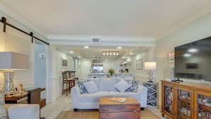A living room with a sofa, coffee table, and wooden cabinet. There is a kitchen in the background with a dining area. - Oceanfront PCB Condo w/ Beach Access & 2 Pools (Panama City Beach)