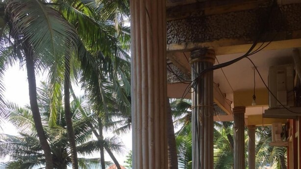 A covered outdoor area with columns, a brick wall, and a seating area with chairs. - Nimmu House (Kumta)