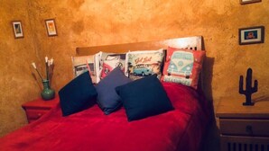Una cama con una colcha roja y varios almohadones, una mesita de noche con un florero, y un cactus decorativo en la pared.