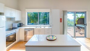 A modern kitchen with white cabinets, a stainless steel dishwasher, and a microwave. There is a window with a view of greenery outside. - Angourie Blue 8 Now Pets considered (Angourie)