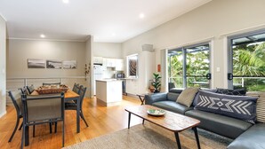 A modern living room with a sofa, coffee table, and dining area. There is a kitchen in the background with white cabinets and a microwave. - Angourie Blue 8 Now Pets considered (Angourie)