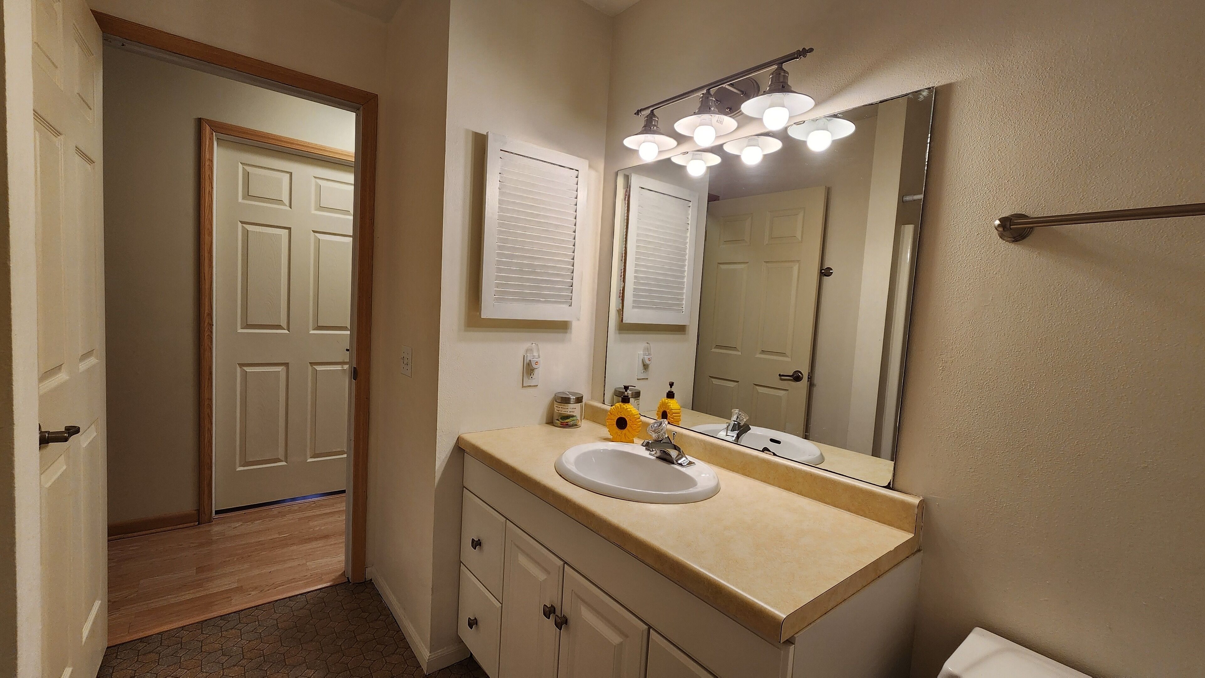 A bathroom with a white sink, vanity, and a large mirror with lights. There are two doors, one leading to another room and the other to the bathroom.