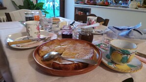 Ein Esstisch mit einem großen Obstkuchen, einem Messer und einem Löffel. Auf dem Tisch befinden sich verschiedene Tassen, ein Glas und eine Vase.