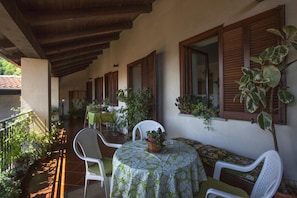 Un espacio de estar al aire libre techado con mesa y sillas, rodeado de plantas en macetas y contraventanas de madera.