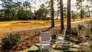 A park with a paved path, white rocking chairs, and a grassy area with trees.