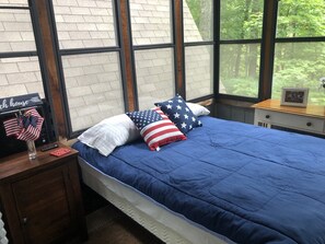 Une chambre avec une grande fenêtre, un lit avec une literie bleue et une table de chevet décorée d’un drapeau américain.