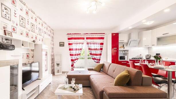 A modern living room with a sofa, TV, and white coffee table. There is a kitchen area with red cabinets and a dining table.