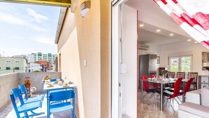 A balcony with blue chairs and a table set for dining, a kitchen area with a dining table and chairs, and a view of buildings in the background.