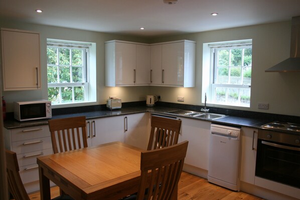 A compact kitchen with white cabinets, a microwave, a dishwasher, and a stove. There are two windows providing natural light and a view of greenery outside.