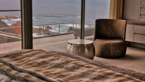 Une chambre moderne dotée d’une grande fenêtre donnant sur l’océan, d’un lit confortable, d’une table d’appoint ronde et d’un fauteuil marron.