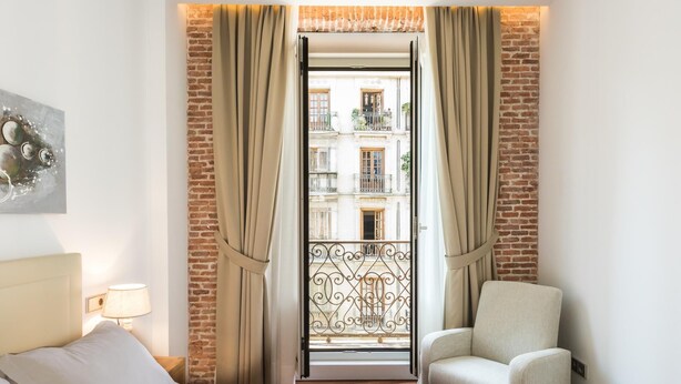 A hotel room with a bed, a chair, a table with a towel, and a view of a building through a window with curtains. - PUERTA DEL SOL CITY CENTER III WITH BALCONY (Madrid)