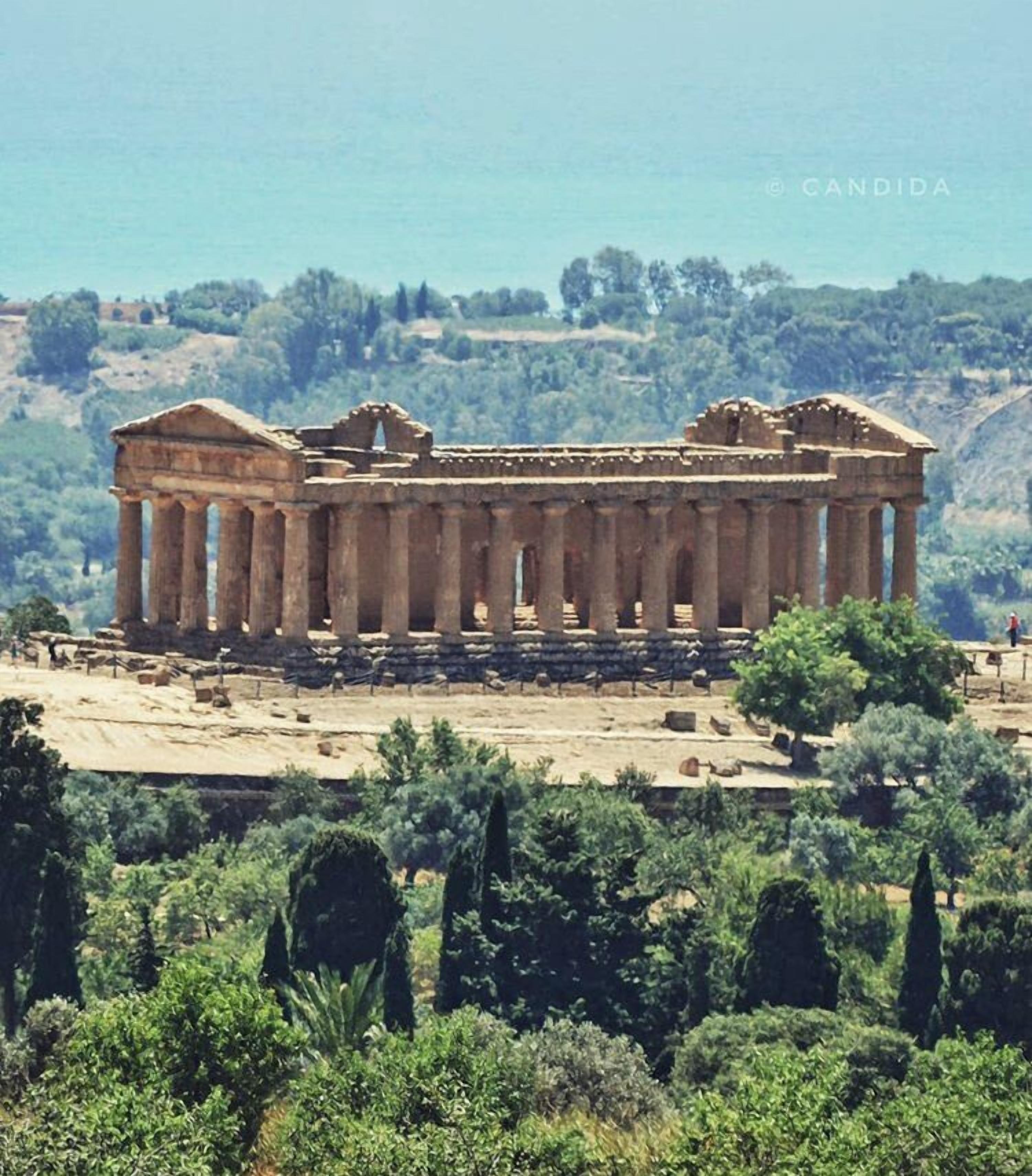 Ein antiker griechischer Tempel mit zahlreichen Säulen und einer flachen Dachkonstruktion, umgeben von Grünflächen und Hügeln im Hintergrund.