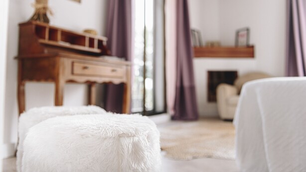 Una habitació acollidora amb un escriptori de fusta, un otomà blanc i esponjós i una cadira blanca.
