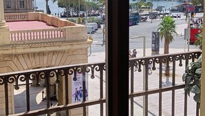 Vista des d'un balcó amb barana metàl·lica, sobre una rambla animada amb cotxes i vianants, una massa d'aigua i edificis, incloent-hi una torre.