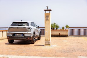 A parked car with a security camera on a post, a paved area with plants, and a railing. - Little Sunshine apartment (Taormina)