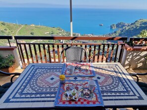 Un balcón con una mesa de baldosas dispuesta para dos personas, con un mantel decorativo y un plato pequeño con comida.