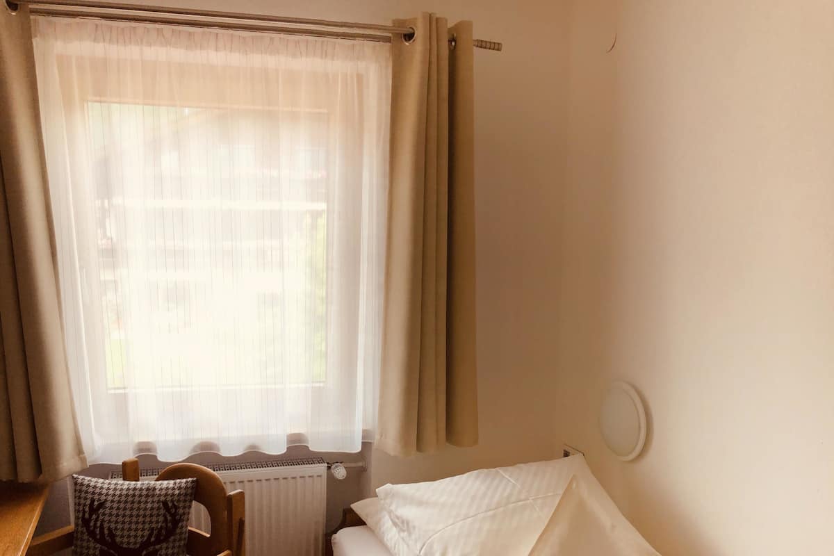 A small, single-bed room with a wooden chair, a radiator, and a window with curtains.
