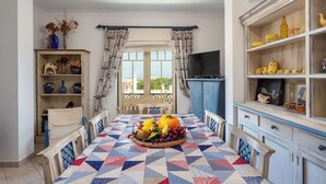 A dining area with a table set for two, a window with curtains, and a shelf with decorative items. - Amazing Duplex w/ Stunning Pool by HostWise (Albufeira)
