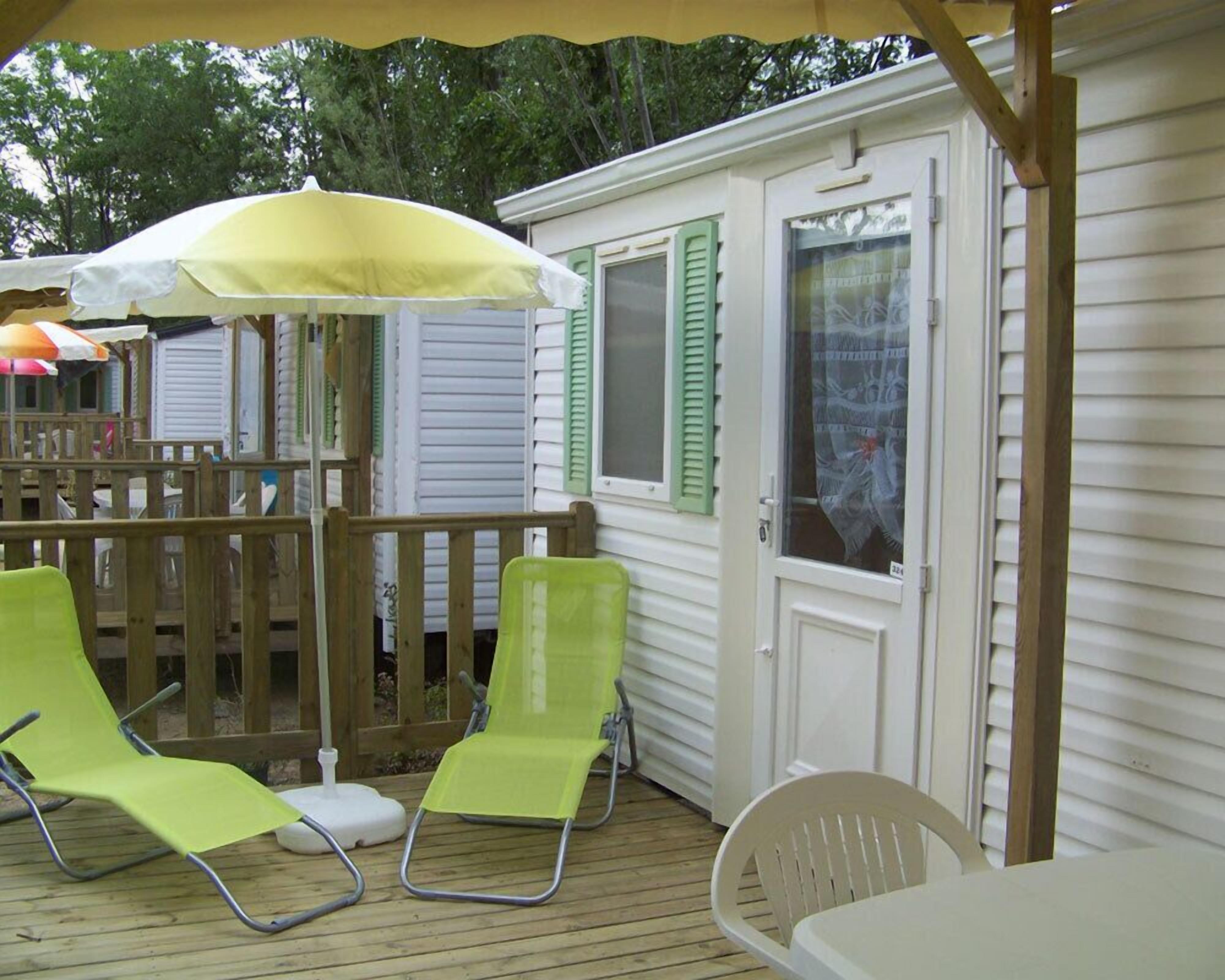 A wooden deck with two green lounge chairs and a white table, a yellow umbrella, and a white door with green shutters.