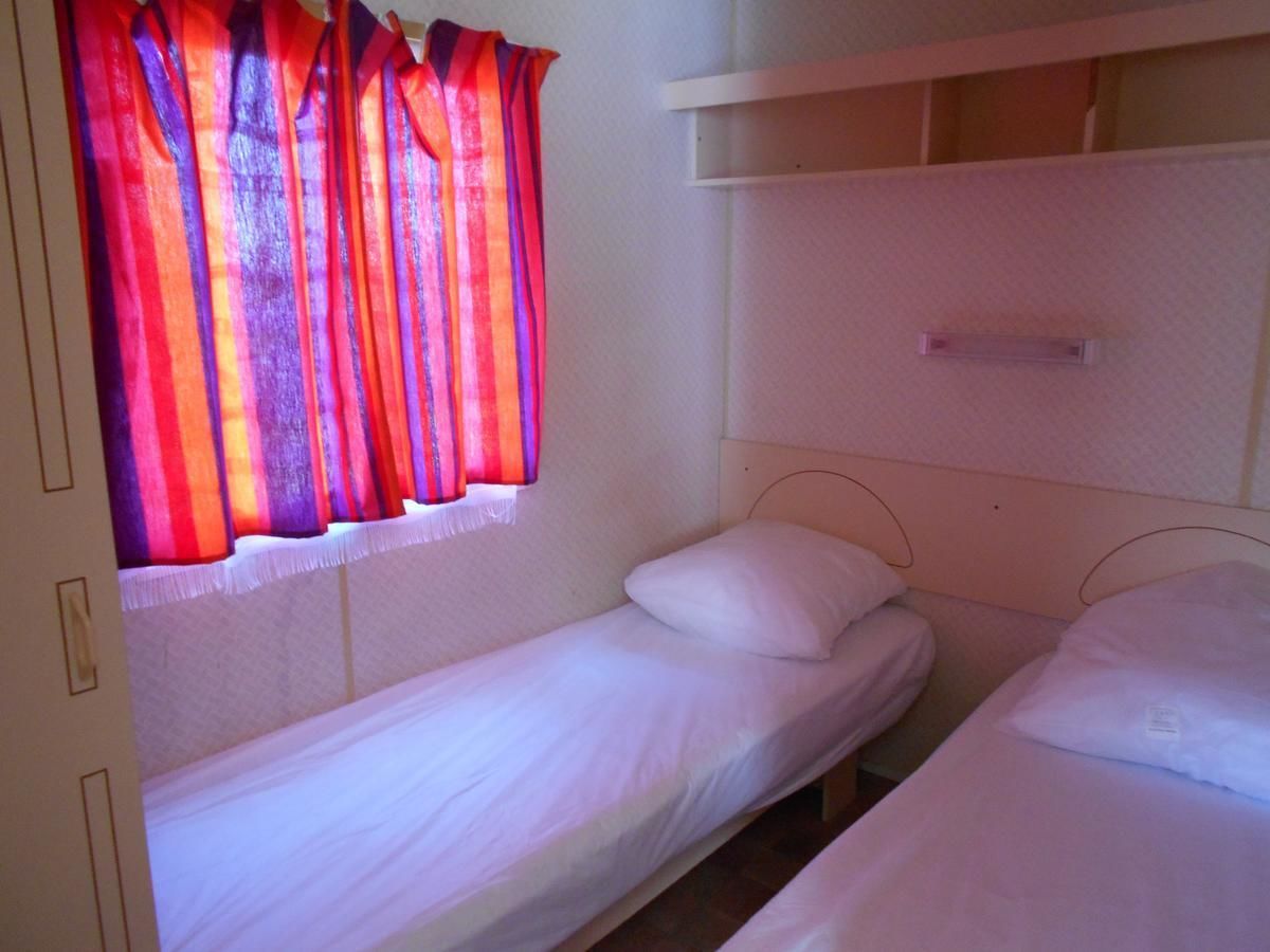 Un petit logement composé d’une seule pièce, comprenant deux lits simples, une fenêtre avec des rideaux rouges et une tête de lit blanche.