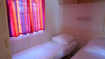 Un petit logement composé d’une seule pièce, comprenant deux lits simples, une fenêtre avec des rideaux rouges et une tête de lit blanche.