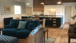 A spacious living area with a high ceiling, exposed beams, and a brick wall. There is a blue sofa, a wicker chair, and a wooden table. A kitchen area is visible in the background with a white countertop and a hanging light fixture. - Number 6 Narrow Water Castle (Warrenpoint)