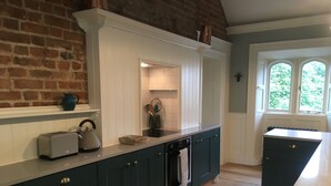 A kitchen with dark cabinetry, a white countertop, a brick wall, and a window with a view of greenery. - Number 6 Narrow Water Castle (Warrenpoint)