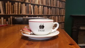 Una taza blanca con el año 1938 sobre un platito, sobre una mesa de madera en una habitación con una estantería y una chimenea.