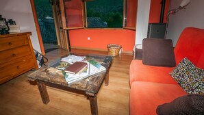 Un salon avec une table basse en verre, un canapé rouge, une commode en bois et une fenêtre donnant sur un paysage verdoyant.