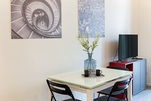 A dining area with a table, chairs, a vase with plants, a television on a cabinet, and two artworks on the wall.