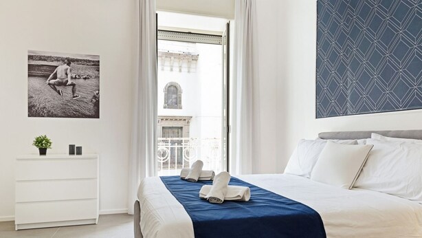 Une chambre d’hôtel avec un lit, une commode blanche, une plante, un tableau encadré et une vue sur un immeuble par la fenêtre.