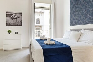 Et hotelværelse med en seng, en hvid kommode, en plante, et indrammet billede og udsigt til en bygning gennem et vindue.
