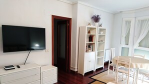 Una sala de estar moderna con un televisor de pantalla plana, muebles blancos, piso de madera y una zona de comedor con mesa y sillas.