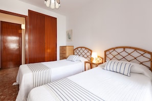 Quarto de hotel com duas camas, cabeceiras de madeira, uma mesa de cabeceira com abajur e um guarda-roupa de madeira.