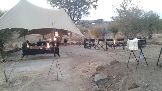A tent with a dining table set up under it, surrounded by chairs and a small fire. - Pop Lamont Camp (Ngoma)