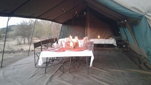 An outdoor dining setup in a tent with tables set for a meal, surrounded by natural landscape. - Pop Lamont Camp (Ngoma)