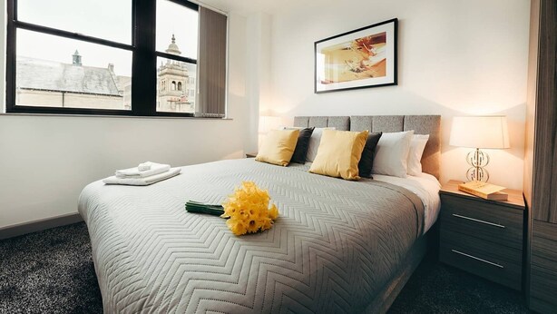 Une chambre d’hôtel avec un grand lit, une table de chevet, une lampe, un tableau encadré au mur et une fenêtre donnant sur des immeubles.