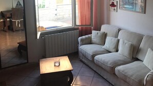A living room with a sofa, a small table, a radiator, and a window with curtains. - THE TERRACE ON THE CREW (Santa Teresa Gallura)
