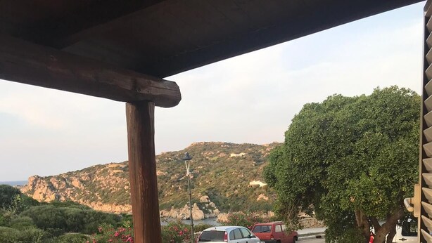 A covered patio with a table and chairs, overlooking a parking area and a hilly landscape. - THE TERRACE ON THE CREW (Santa Teresa Gallura)