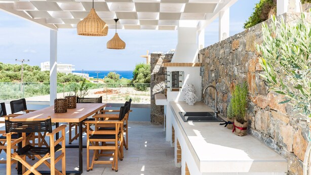 En udendørs terrasse med et træbord og stole, en stenvæg med grønne planter, og udsigt til havet.