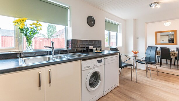 A modern kitchen with a washing machine, a dining area with a glass table, and a clock on the wall.
