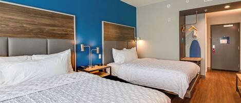 Quarto de hotel com duas camas, cabeceira de madeira, parede azul e porta com espelho e cabides.