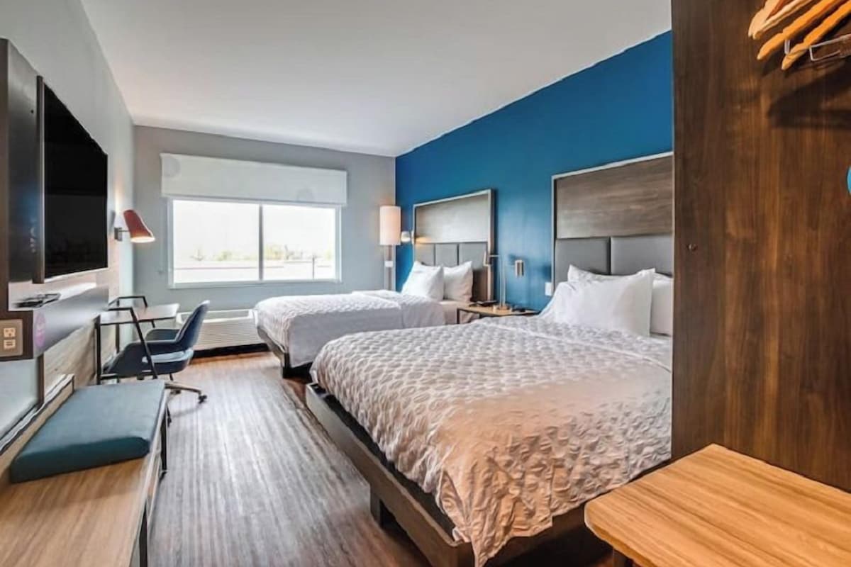 A hotel room with two beds, a desk, a chair, a TV, and a large window.
