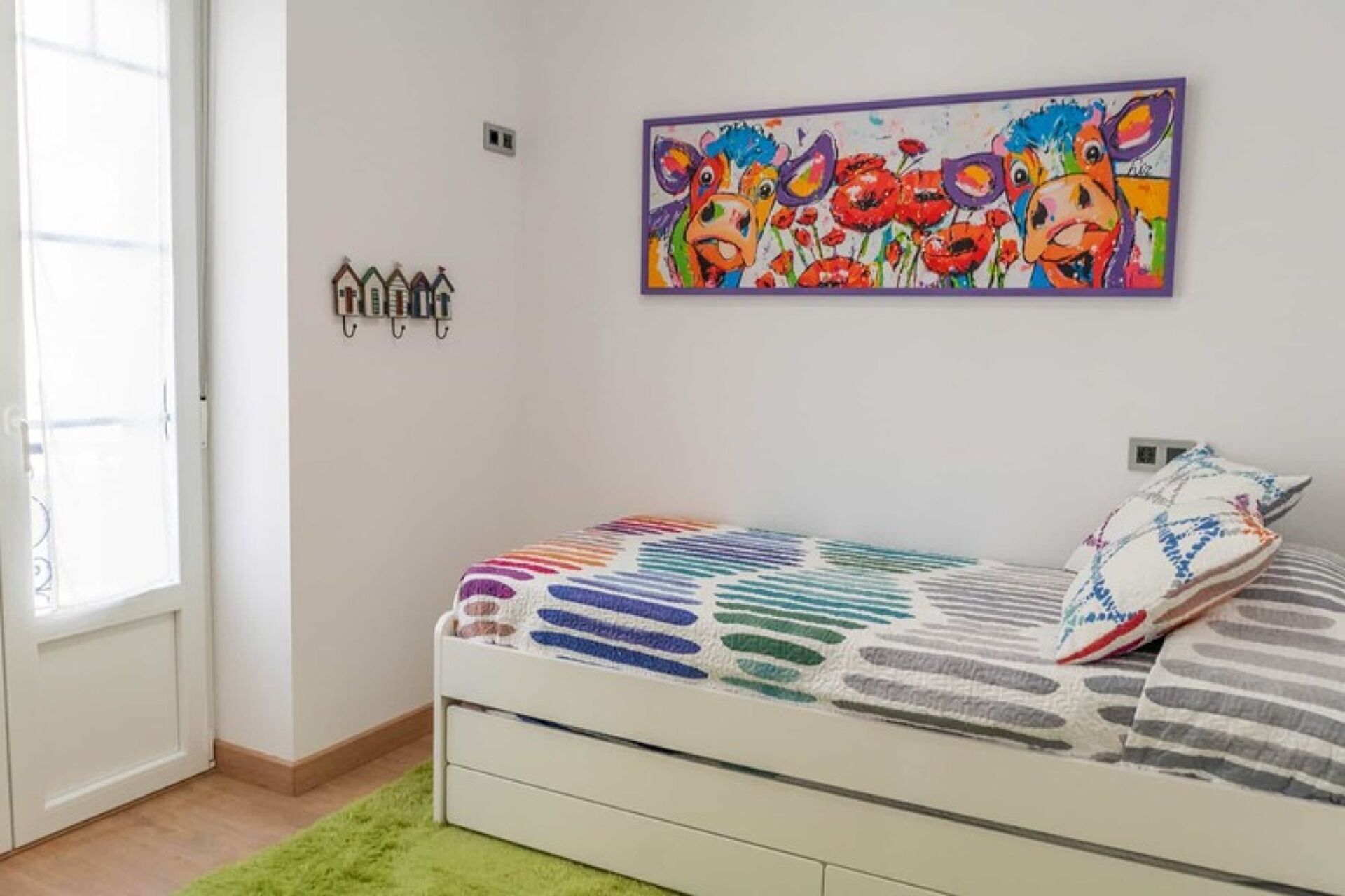Un dormitori amb un llit, un quadre colorit, una finestra i una porta.