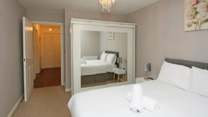 A hotel room with a large bed, a bedside table with a lamp, a mirror, and a door leading to another room. - Autumn Heights Apartments (Telford)