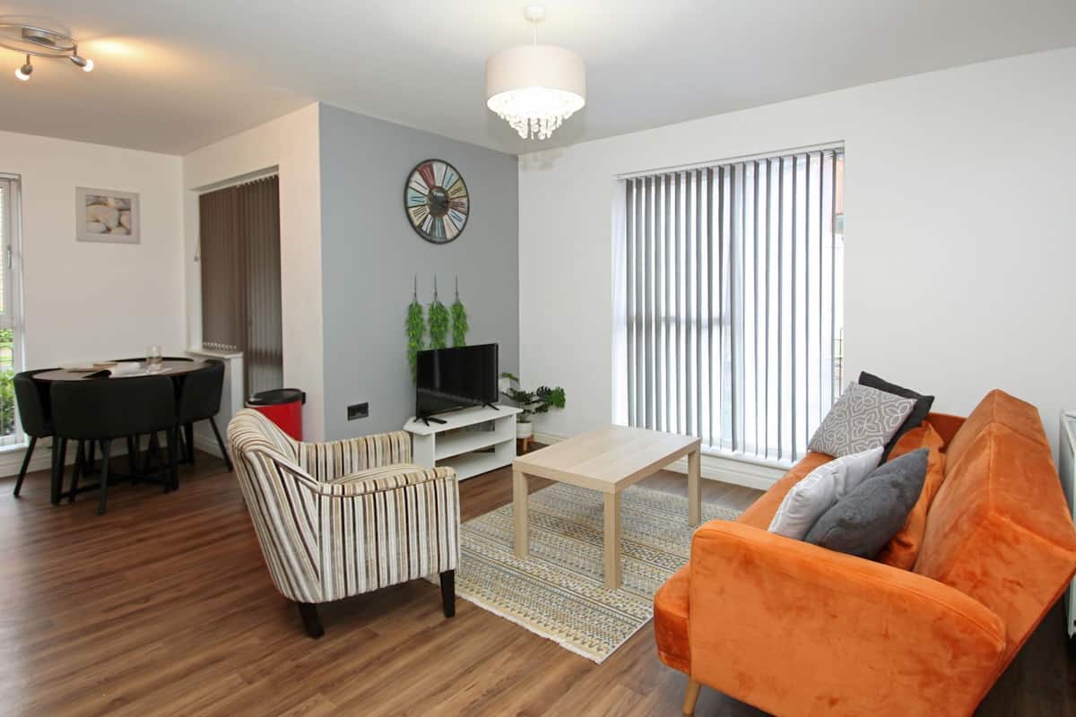 A modern living room with a striped armchair, a wooden coffee table, an orange sofa, a TV on a white stand, a clock on the wall, and a window with blinds.