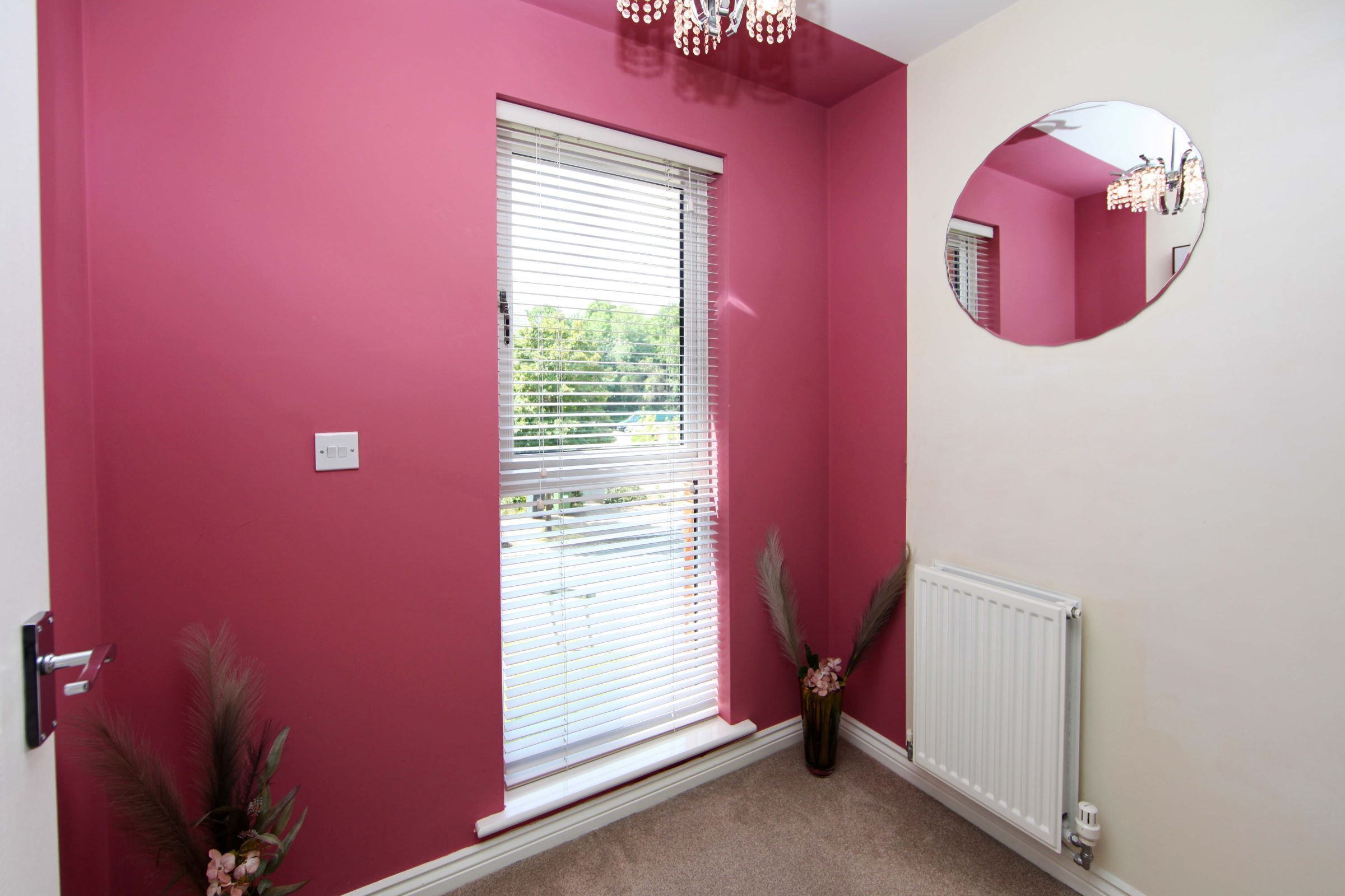 Une pièce avec un mur rouge, un radiateur blanc, une fenêtre avec des stores, un miroir et une porte.